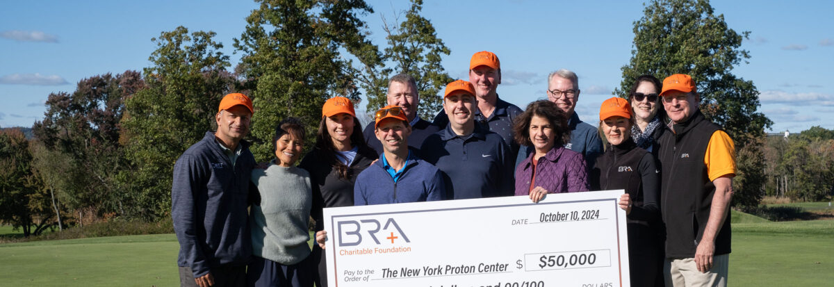 A group of men and women gathered outdoors on a green lawn, holding a large check for a $50,000 donation to the New York Proton Center.