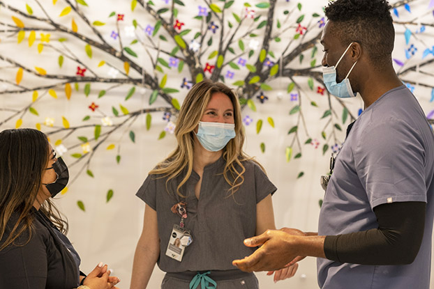 Three nurses wearing masks facing each other in front of a mural speaking to each other jovially.
